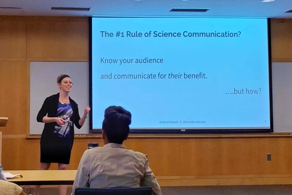 Stephanie speaking at the front of a room in front of a presentation screen
